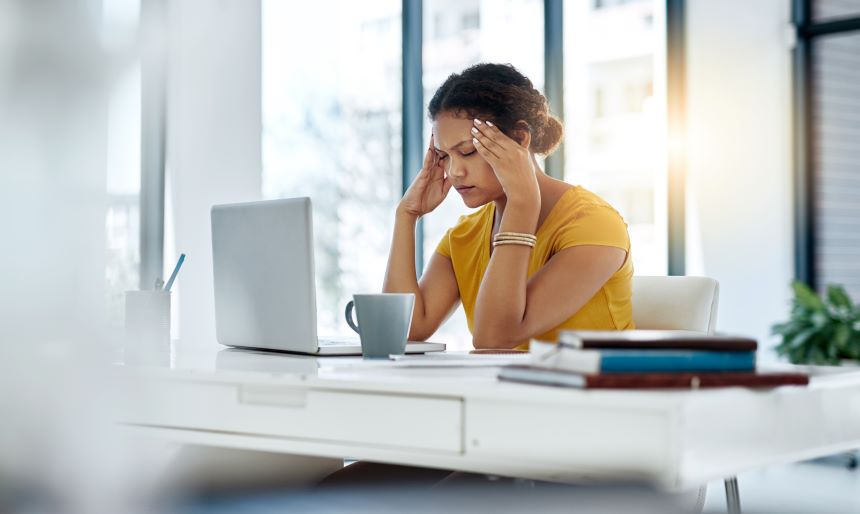 A young professional woman sits at her desk at work holding her head from a headache or migraine.