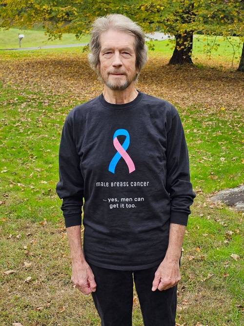 Alan Smith standing outside wearing a black t-shirt with a multicolored cancer ribbon that says “Male breast cancer. Yes, men can get it too.”