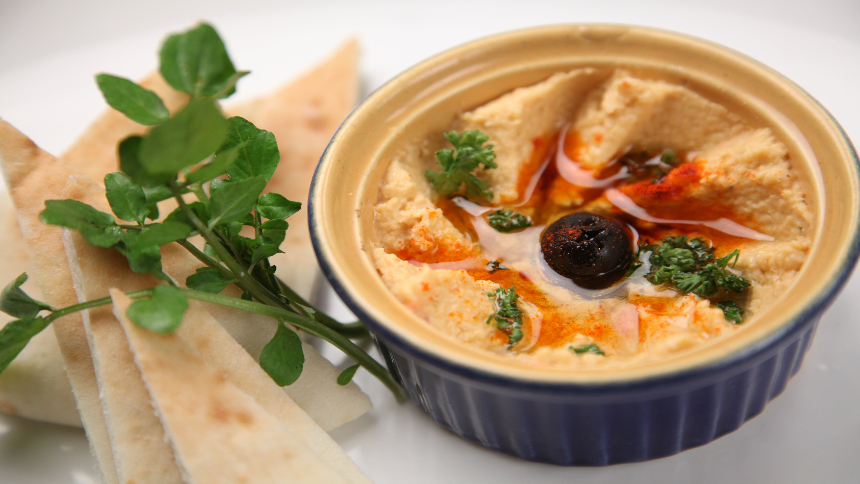 A bowl of hummus garnished with paprika, olive oil, a whole black olive, sliced onions, and parsley, served with pita bread and fresh watercress on the side.