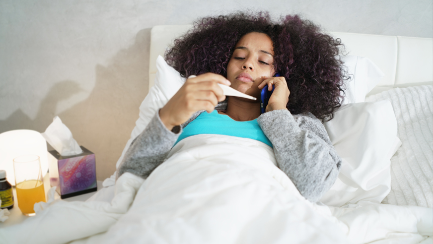 Sick high school girl lies in bed, reading a thermometer with a phone by her ear