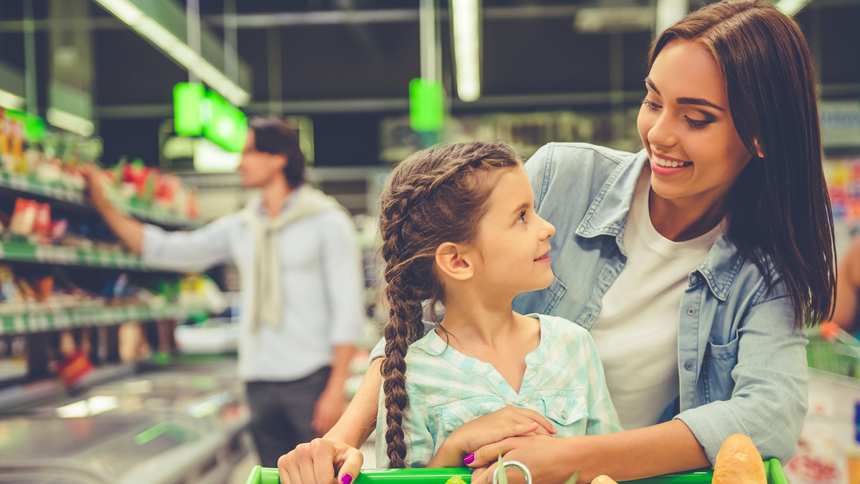 A mom shops with her daughter in a grocery store with produce in the cart