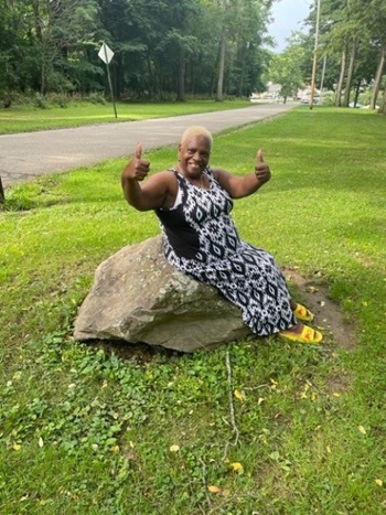 Barbara Brickhouse, 68, Beacon, New York. Barbara is a two-time cancer survivor. She is sitting on a rock outside in a park in the summer.