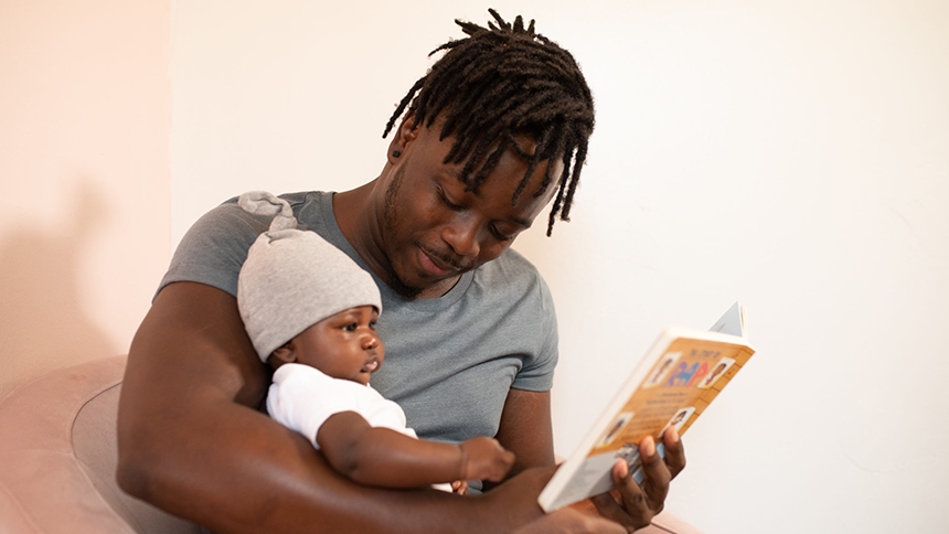 A father which his young child sitting in his lap; he is holding a book and reading to the child