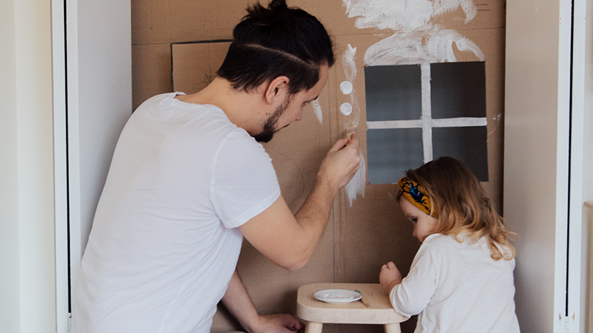 A father and daughter painting a pretend house, the house is made of cardboard.