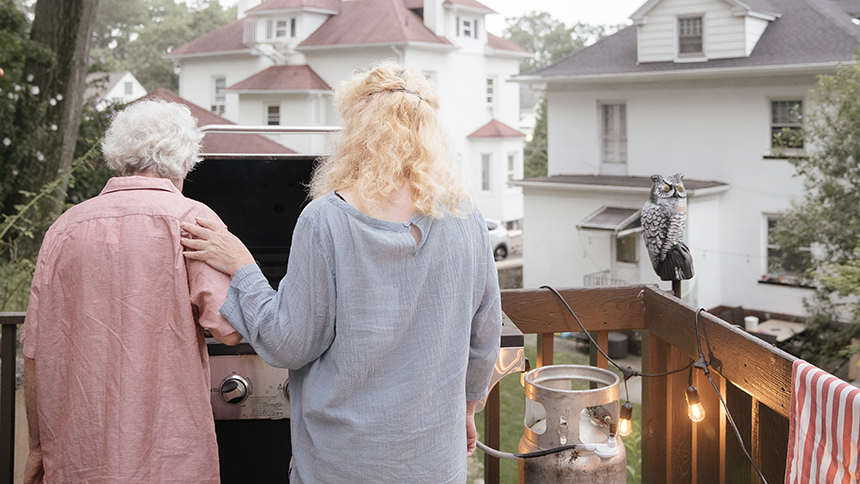 An elderly mother and her daughter barbecuing outside on a porch. Their backs are to the camera and they're facing the grill.