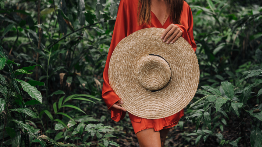 A photo of a woman walking through a tropical forest holding a woven sunhat in front of her stomach. She is wearing an orange sundress. 