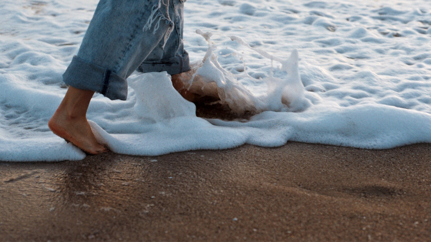 A photo of a person wearing jeans walking through a foamy wave on the beach, showing the person only the knees down.