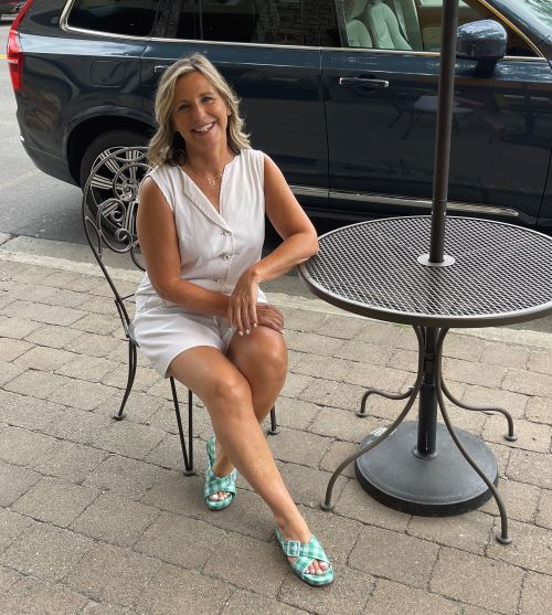 Gina Lacey, a 53-year-old woman from New Milford, Connecticut who had breast cancer, is sitting outside at a table wearing a summer outfit.