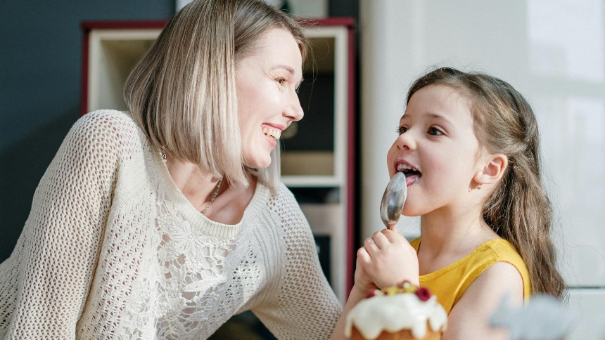 Woman in white knit sweater smiling while a little girls licks icing off a spoon