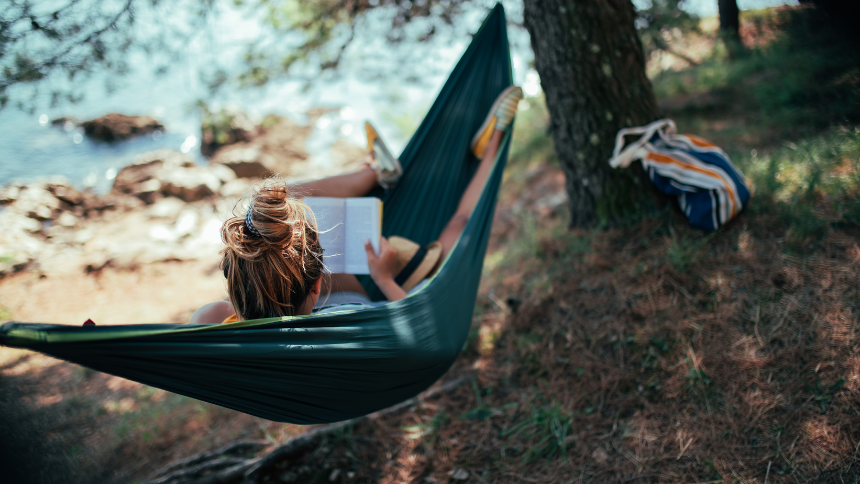 Young woman in a hammock and reading a book outside near a lake. Reading has many brain-boosting benefits and is good for your mental and physical health.