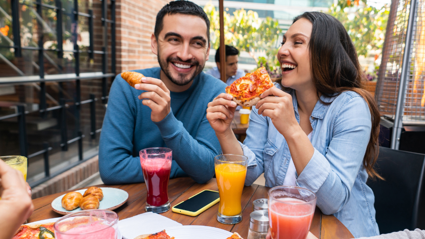 A Caucasian man and woman sitting at an outdoor table eating pizza and enjoying time with friends because they are taking care of their digestive health.  