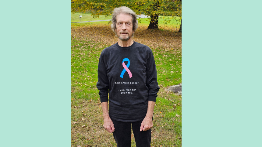 Alan Smith standing outside wearing a black t-shirt with a multicolored cancer ribbon that says “Male breast cancer. Yes, men can get it too.”