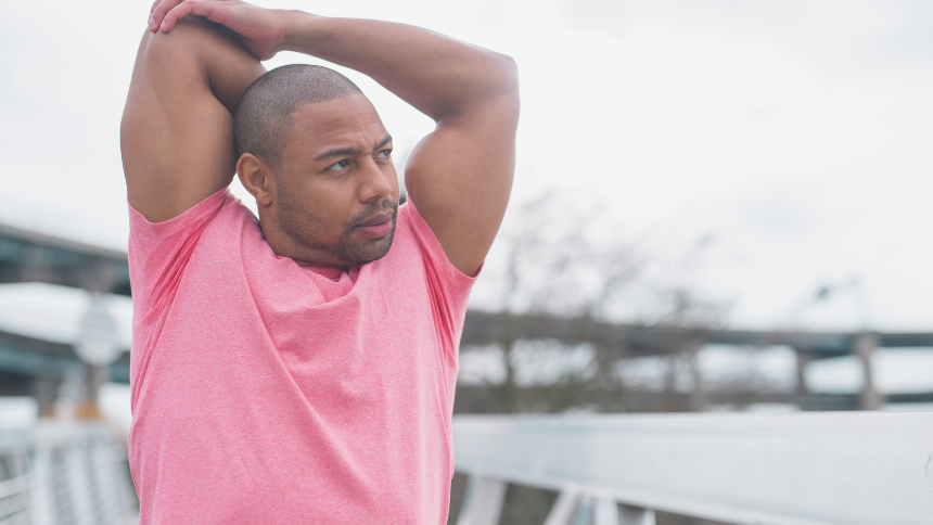 Man wearing a pink shirt stretching his tricep outside near a body of water and a bridge.