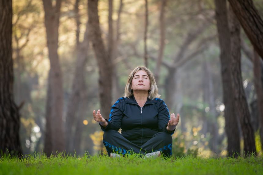 A middle-aged woman sitting cross-legged on grass and meditating near tall trees.