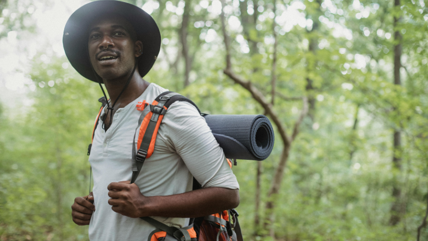 Adult man wearing a wide-brimmed hat and backpack with yoga mat hiking through the woods.