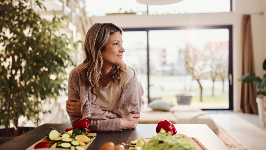 Young smiling woman daydreaming while spending time in dining room and preparing a healthy meal of fruits and vegetables. 