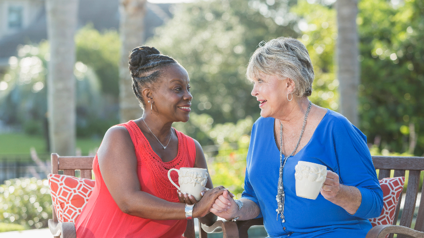 Two senior multi-ethnic women, one Caucasian and the other African American, sitting on a porch enjoying each other’s company. They are facing each other, smiling, talking and drinking coffee. Ovarian cancer can happen at any age but usually occurs when women are in their 50s, 60s and 70s.