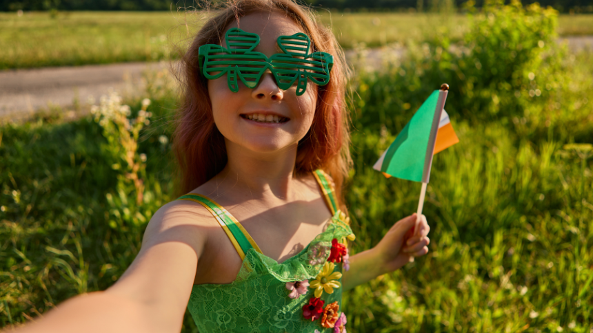 A young girl celebrating St. Patrick’s Day, wearing shamrock glasses, a green sundress with flower decals and waving a small, Irish flag.