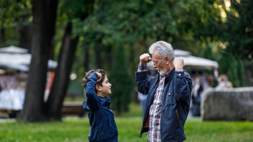 An older man with a beard is enjoying the time with his grandson while playing in a grassy public park.