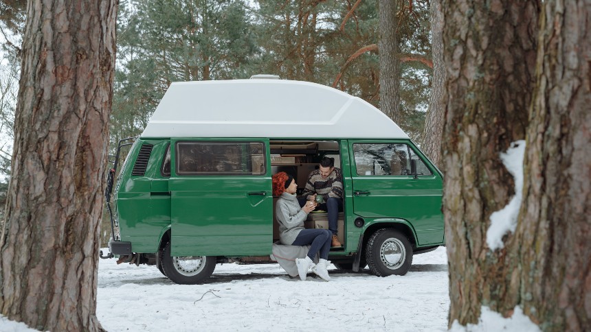 A man and woman standing outside of a green-colored RV in a snowy forest. 