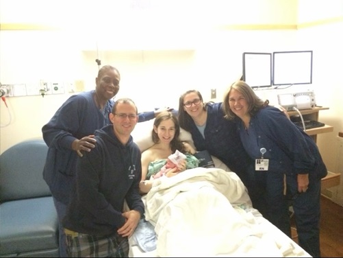 A mom holds her newborn in a hospital bed surrounded by her nurse, midwife and others.