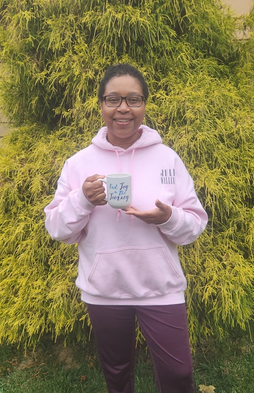 Leonie Roberts, a Norwalk Hospital breast cancer patient, standing outside, wearing a pink sweatshirt and holding a mug that says, “Find joy in the journey”. 