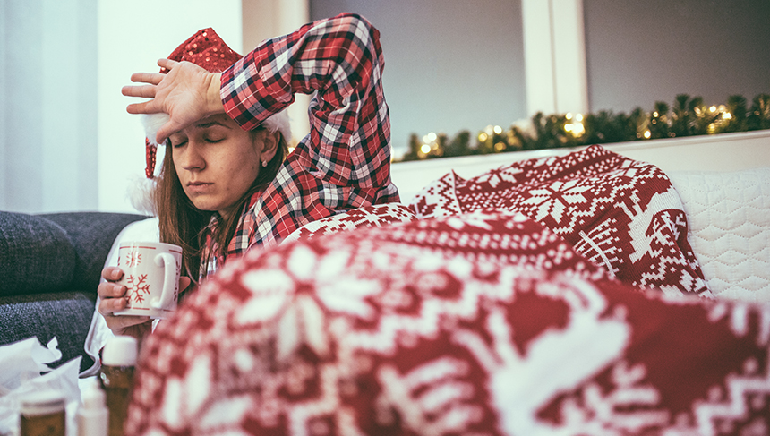 Sick woman in holiday pajamas lays under blankets and checks her temperature. 
