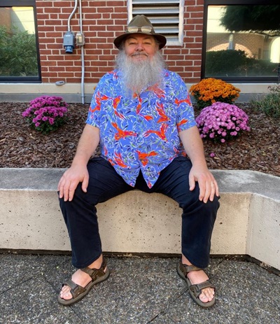 Clarence Shrack, a 61-year-old man from New Milford, CT is sitting outside a garden wearing a wide-brimmed hat and floral shirt. 
