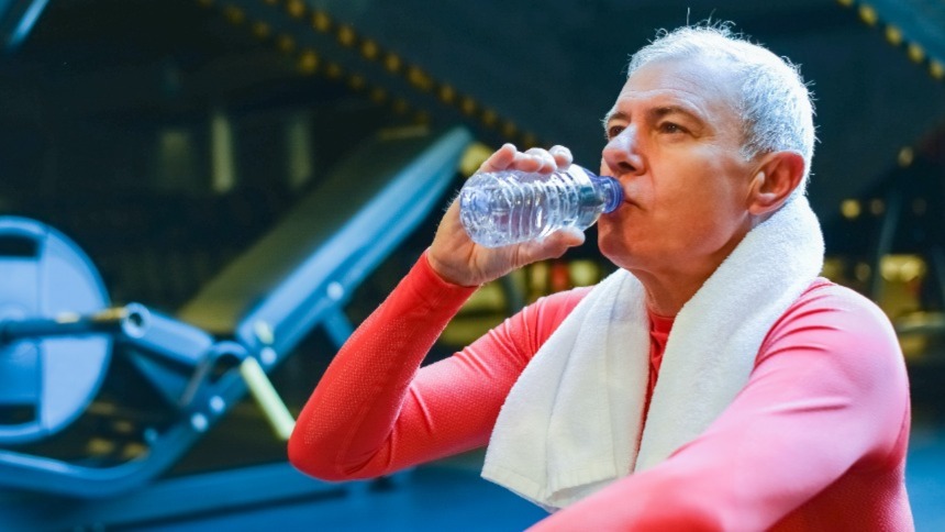 An older Caucasian man with a white towel around his neck sitting on an exercise machine in the gym drinking a bottle of water.