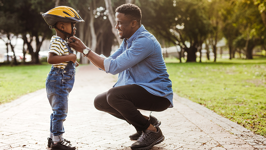 A father fastens a bike helmet on his young son in a park.