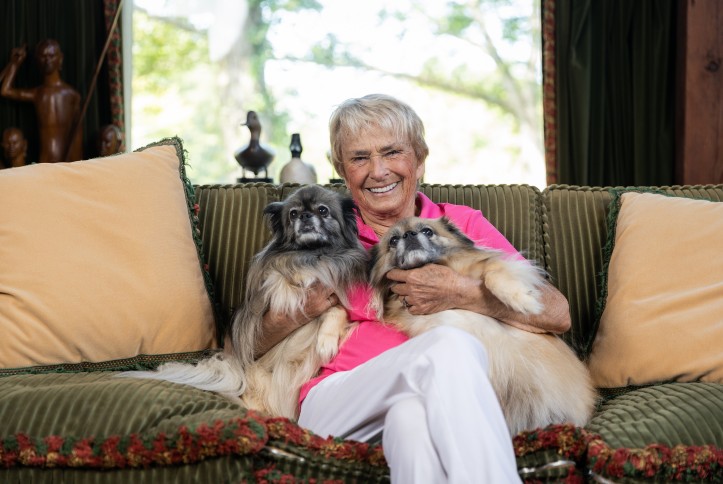 Gayle Bontecou, wearing a pink shirt and white pants, sits on a couch holding two dogs.