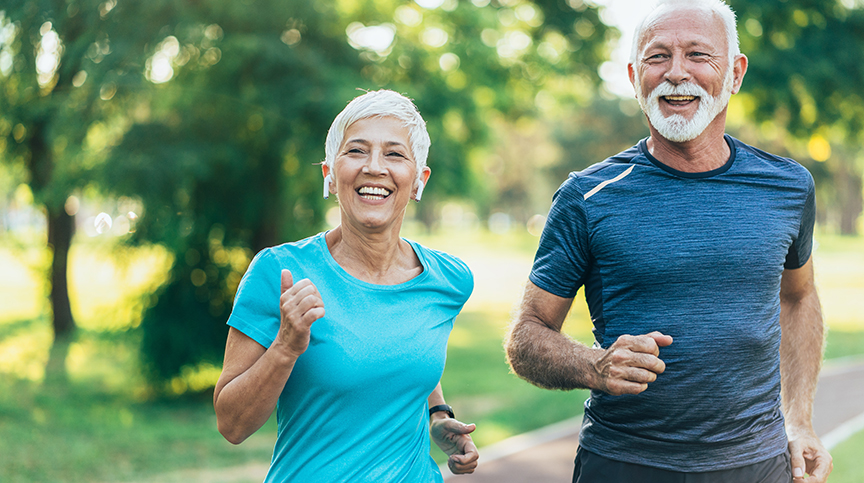  An active and fit older couple go for a speed walk outdoors against a green landscape.