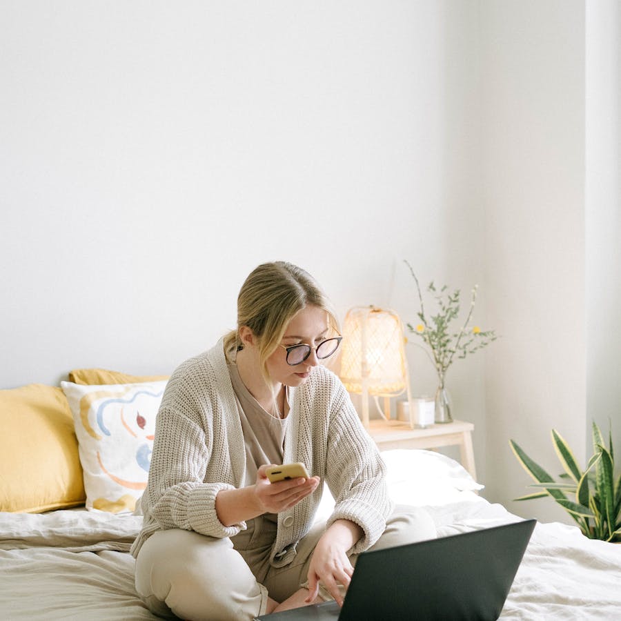 photo of woman sitting on bed while using black laptop