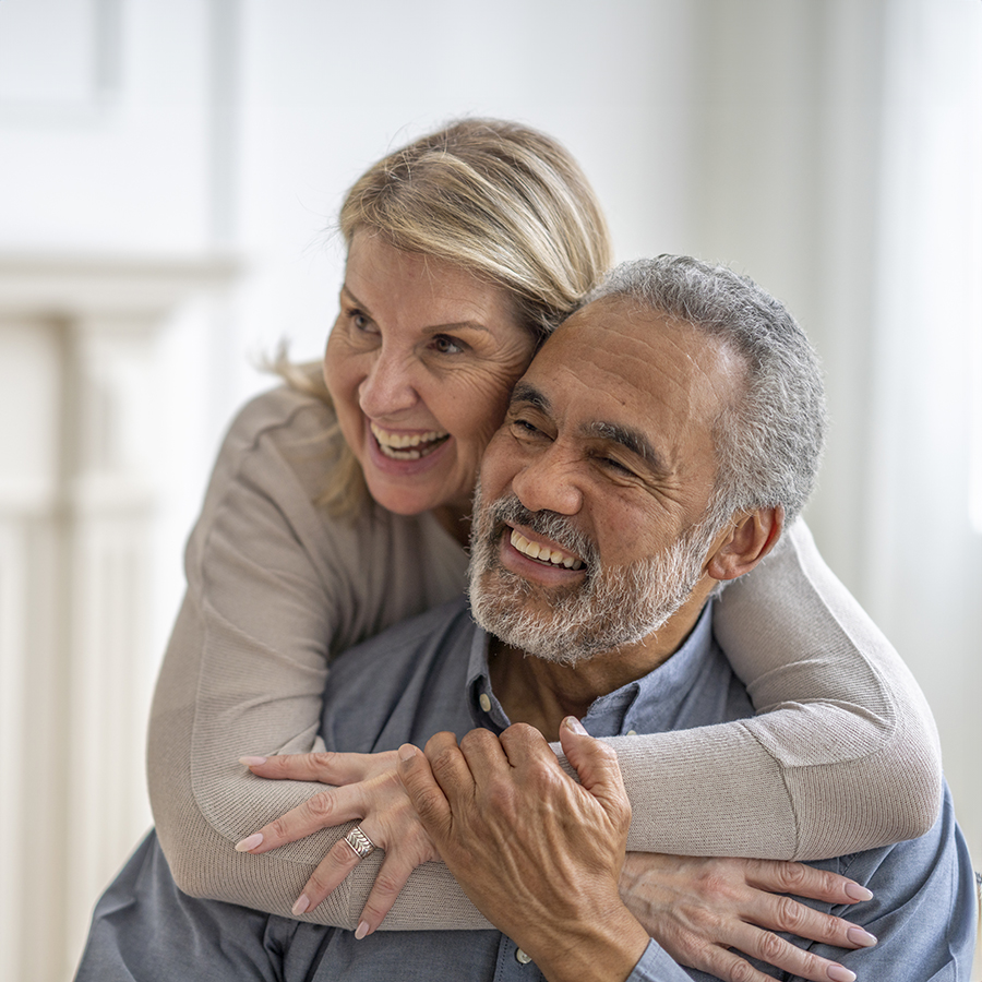 A bi-racial senior couple pose affectionately for a portrait in their living room. The husband is seated on the sofa with his wife standing behind him and wrapping her arms around him in a hug. They are dressed casually and smiling.