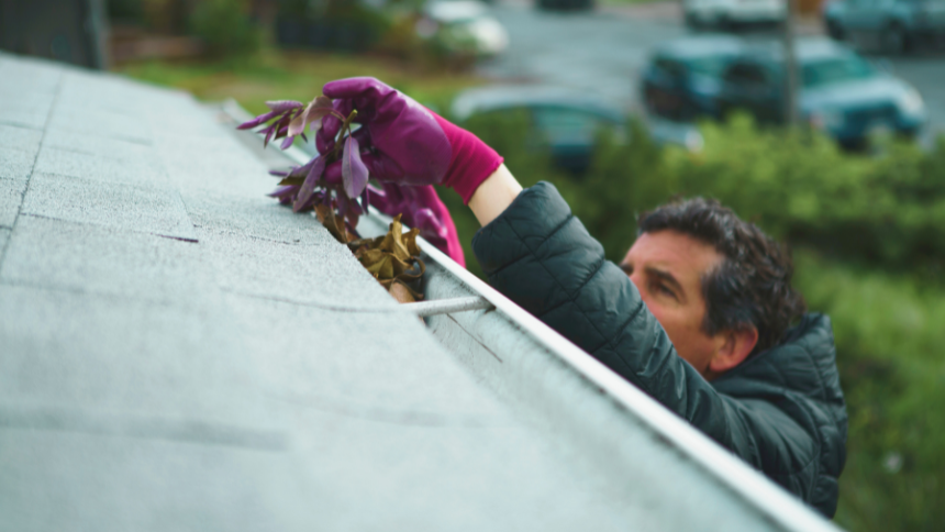 A man wearing a green jacket and red gloves is cleaning out the gutters on his roof. 