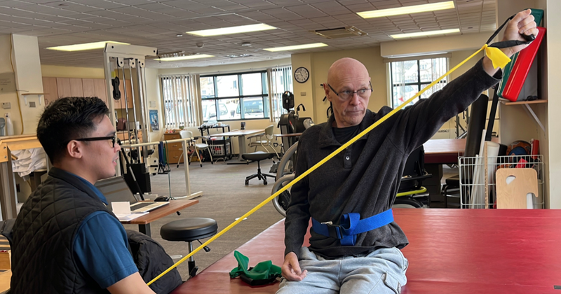 Michael sitting down doing physical therapy. He is using a stretch band to do arm extension exercises while being assisted by his physical therapist.