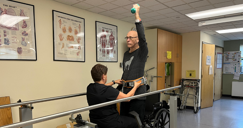 Michael doing physical therapy. He is raising his arm with a dumbbell in hand while being assisted by his physical therapist.