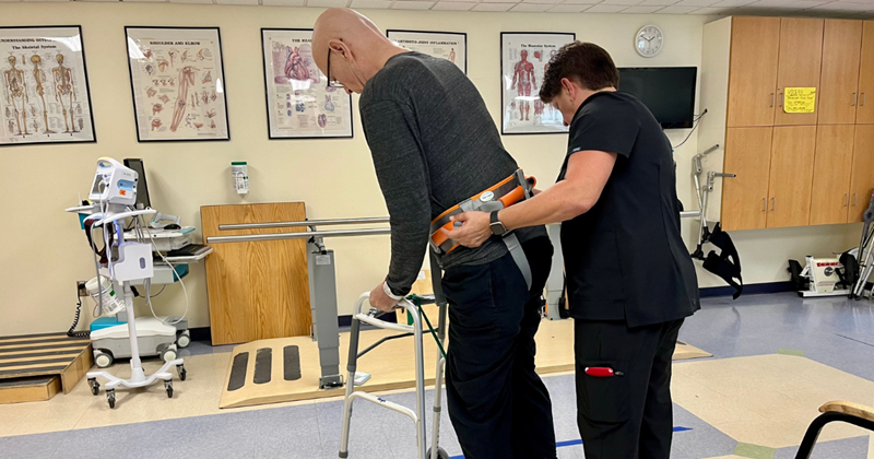 title="Michael doing physical therapy. He is walking with his walker while being assisted by his physical therapist."
