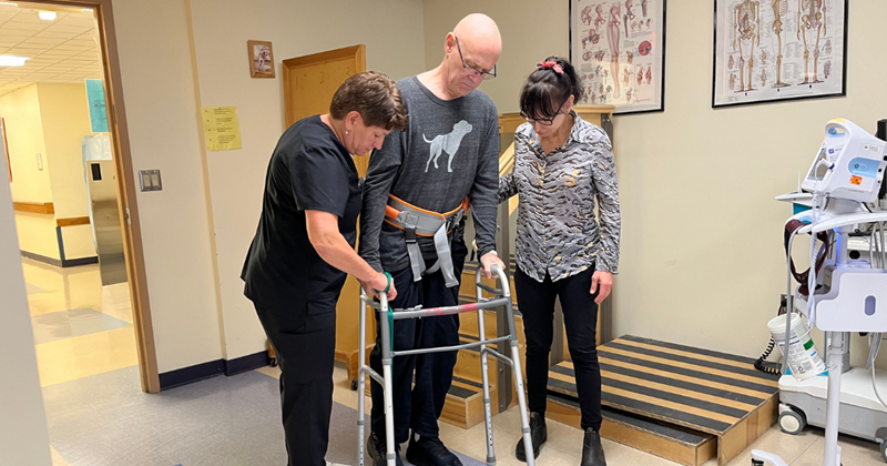 Michael doing physical therapy. He is walking with his walker while being assisted by his physical therapist and Gloria.