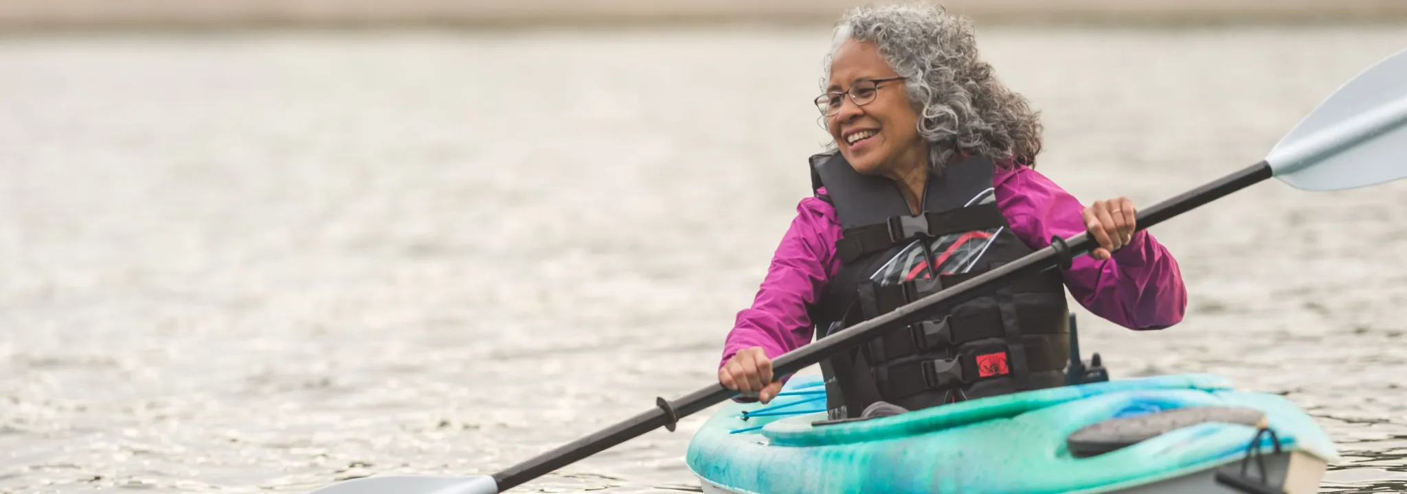 Vibrant senior woman enjoys a beautiful Oregon evening kayaking on the coast. There are trees and a sandbar in the distance behind her.