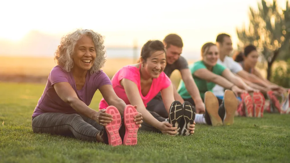 A group of adults attending a fitness class outdoors are doing leg stretches. The participants are arranged in a line. The focus is on a mature woman who is smiling toward the camera.