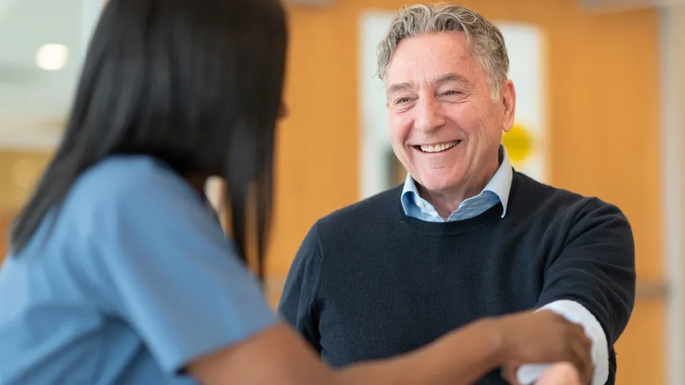 Female nurse is doing a routine check-up with her male patient. The two are seated at a table together. The patient is smiling warmly at his care provider. The nurse is talking.