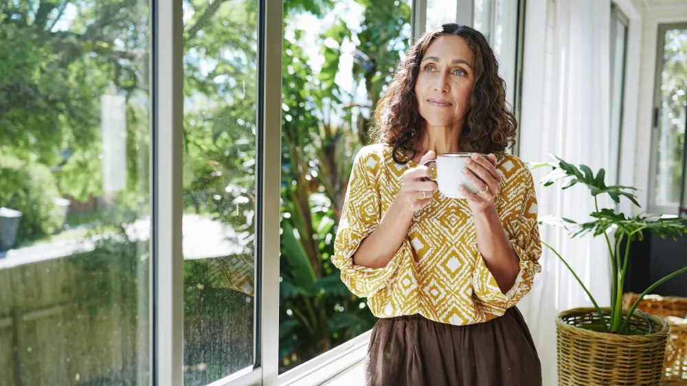 Content mature woman standing by a window at home and looking out at the view while enjoying a cup of coffee in the morning