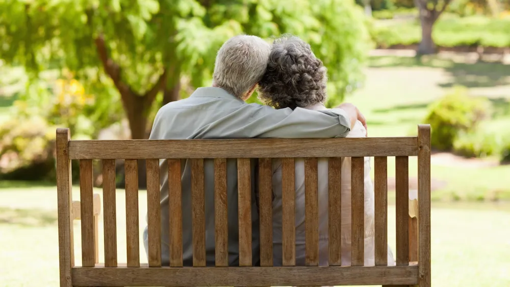 An older couple sitting on a bench with their heads together. The man has his arm around the woman. It is a nice day and they seem to be contemplative.