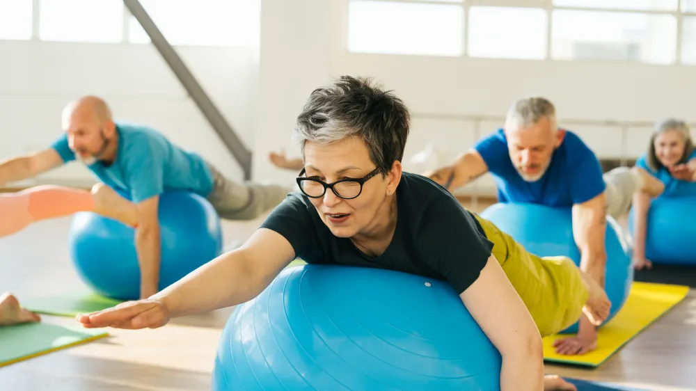 Adults of different ages and ethnicities laying on an exercise ball with an arm extended, stretching their back, which can help to prevent spinal stenosis or lessen symptoms from this common back condition.
