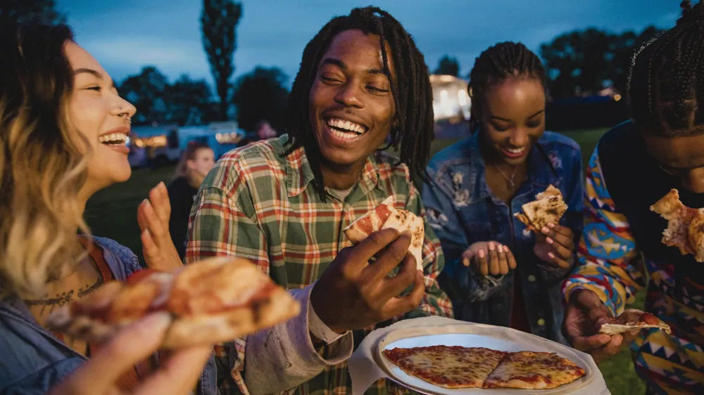 A young group of friends laughing and sharing pizza at a music festival.