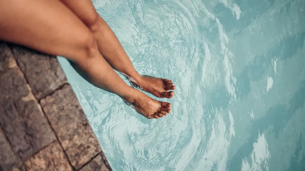 A person sitting poolside with their legs in the water.