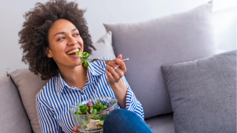 Hero Closeup of a smiling Black woman eating a salad in the livingroom